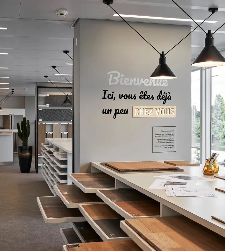 Photo du showroom Kaufman Broad de Courbevoie avec différentes palettes de matériaux pour personnaliser votre futur logement et la phrase inscrite sur un mur :
"Bienvenue
Ici, Vous êtes déjà un peu chez vous"