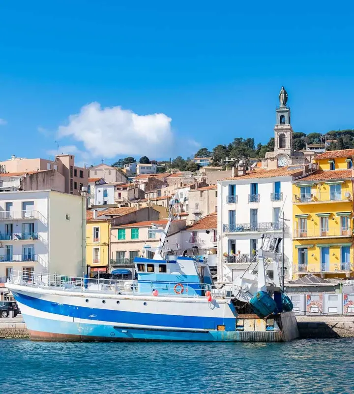 Vue pittoresque d’un port méditerranéen animé, avec un bateau de pêche amarré au premier plan. En arrière-plan, des façades colorées typiques du sud de la France s’élèvent en étages le long de la colline, dominées par un clocher d’église. L’ensemble évoque une atmosphère authentique, conviviale et ensoleillée, idéale pour des projets immobiliers en bord de mer ou dans des quartiers à fort potentiel touristique.