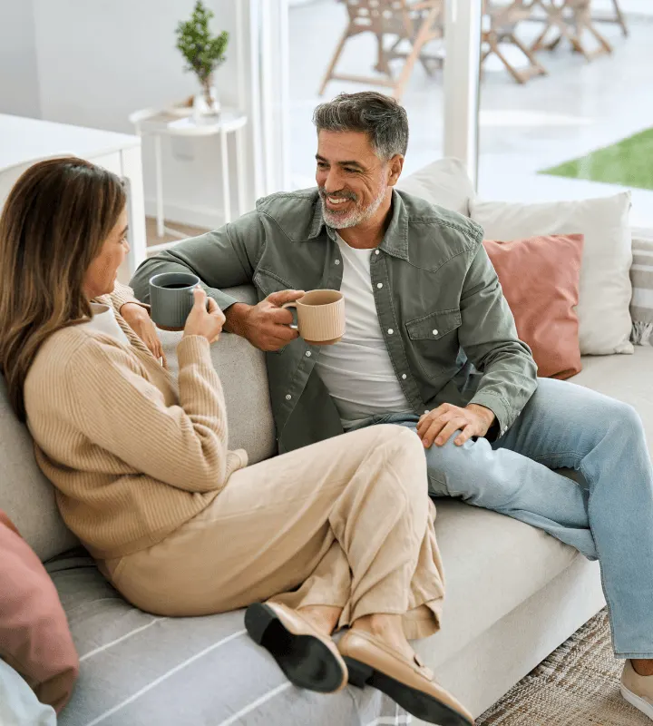 Un couple est assis confortablement sur un canapé, l'homme avec une barbe poivre et sel et une chemise kaki, et la femme aux longs cheveux bruns dans un ensemble beige. Ils se sourient en sirotant du café, transmettant un sentiment de bien-être, de convivialité et de sérénité.