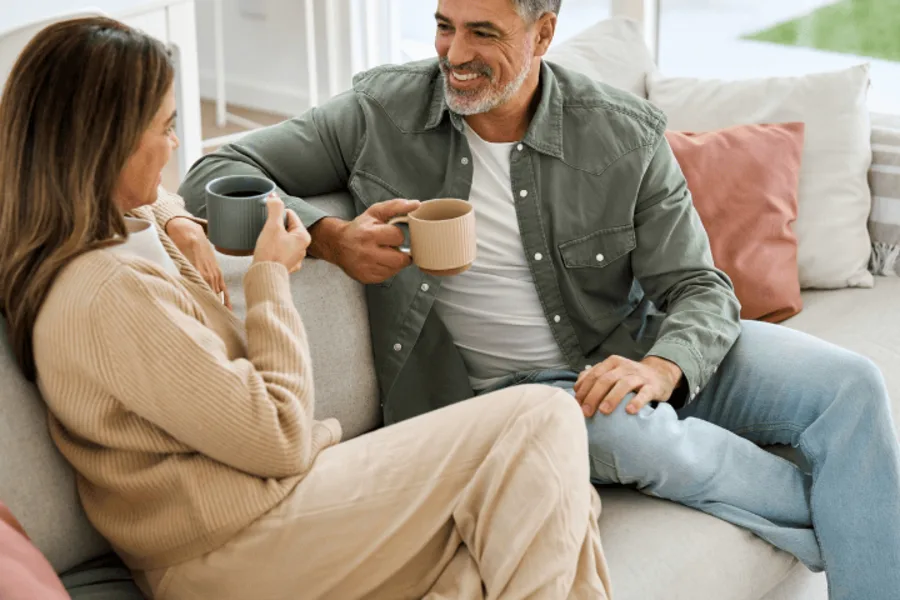 Un couple est assis confortablement sur un canapé, l'homme avec une barbe poivre et sel et une chemise kaki, et la femme aux longs cheveux bruns dans un ensemble beige. Ils se sourient en sirotant du café, transmettant un sentiment de bien-être, de convivialité et de sérénité.
