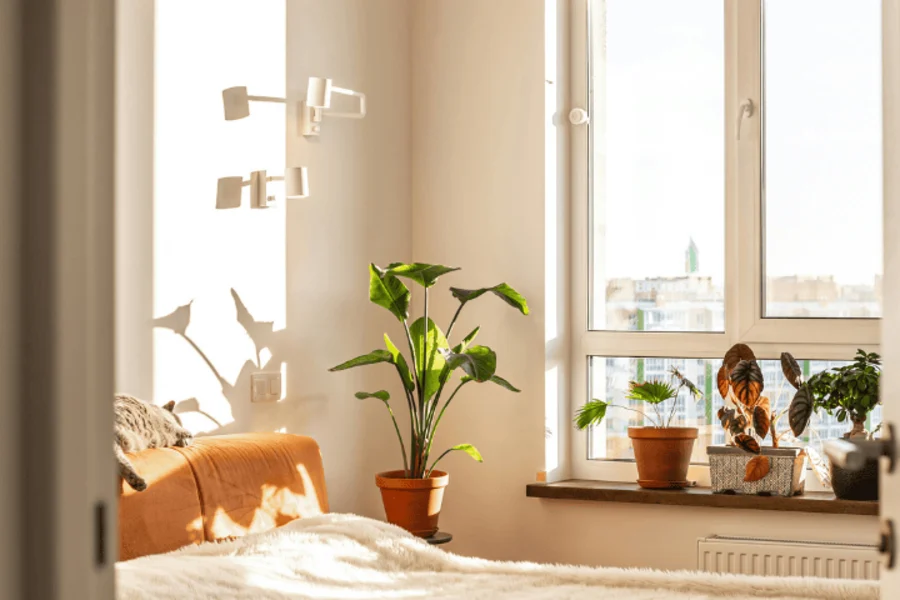 Chambre moderne baignée de lumière naturelle, décorée dans des tons clairs et chaleureux. Un grand lit recouvert d’une couverture beige est placé près d’une grande fenêtre laissant entrer le soleil. Quelques plantes vertes et pots en terre cuite apportent une touche naturelle et apaisante. L’ambiance générale est douce, cosy et minimaliste
