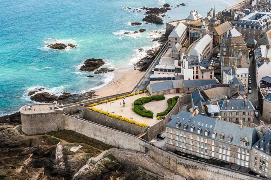 Vue aérienne d’un quartier historique en bord de mer, mettant en valeur une architecture traditionnelle avec des toits en ardoise, entourée de remparts en pierre. L’ensemble est situé face à l’océan, avec une plage de sable fin et des rochers. La présence d’un fort sur un îlot en arrière-plan renforce le caractère patrimonial du lieu. Cette image reflète un cadre de vie authentique et prestigieux, entre patrimoine architectural et panorama maritime.